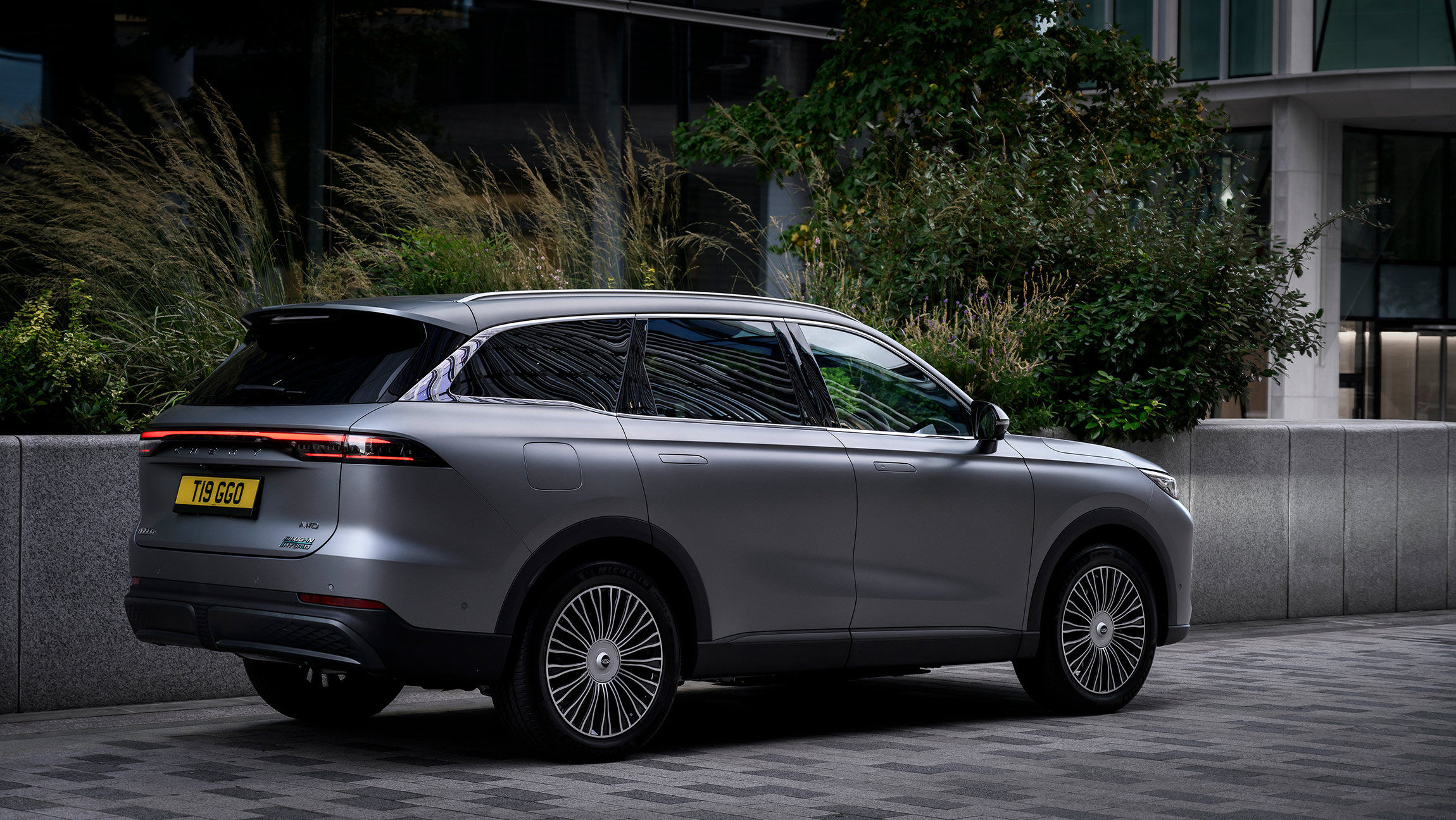 Chery Tiggo 9 CSH SUV rear  view in matte grey finish, parked on a modern city street with greenery in the background.