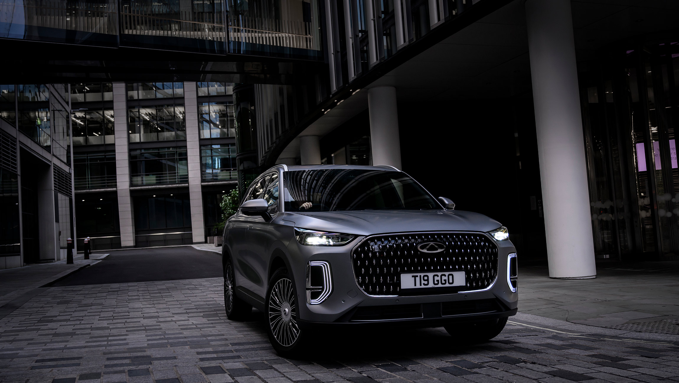 Chery Tiggo 9 CSH SUV in matte grey finish, parked on a modern cobblestone street surrounded by glass-fronted office buildings